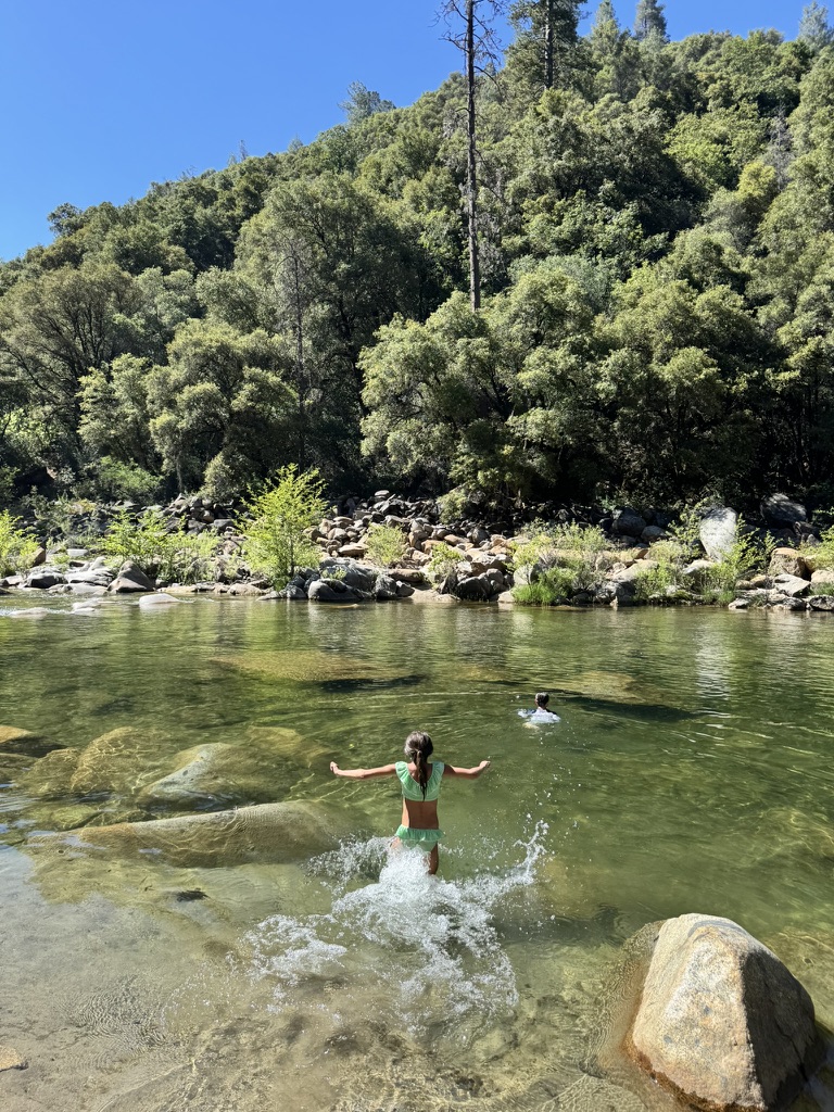 A child wading into the clear green water of the Yuba River, granite boulders and oak forest behind.