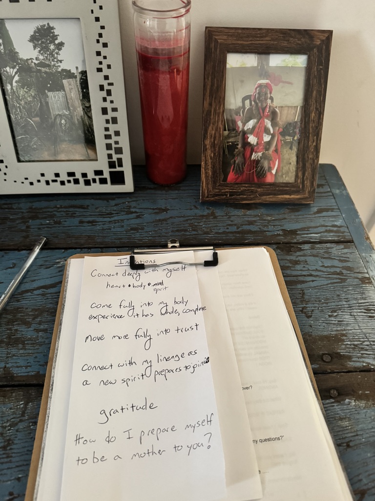 Handwritten intentions on cream paper resting on a clipboard, beside a red candle and a framed photograph of a Bwiti elder.