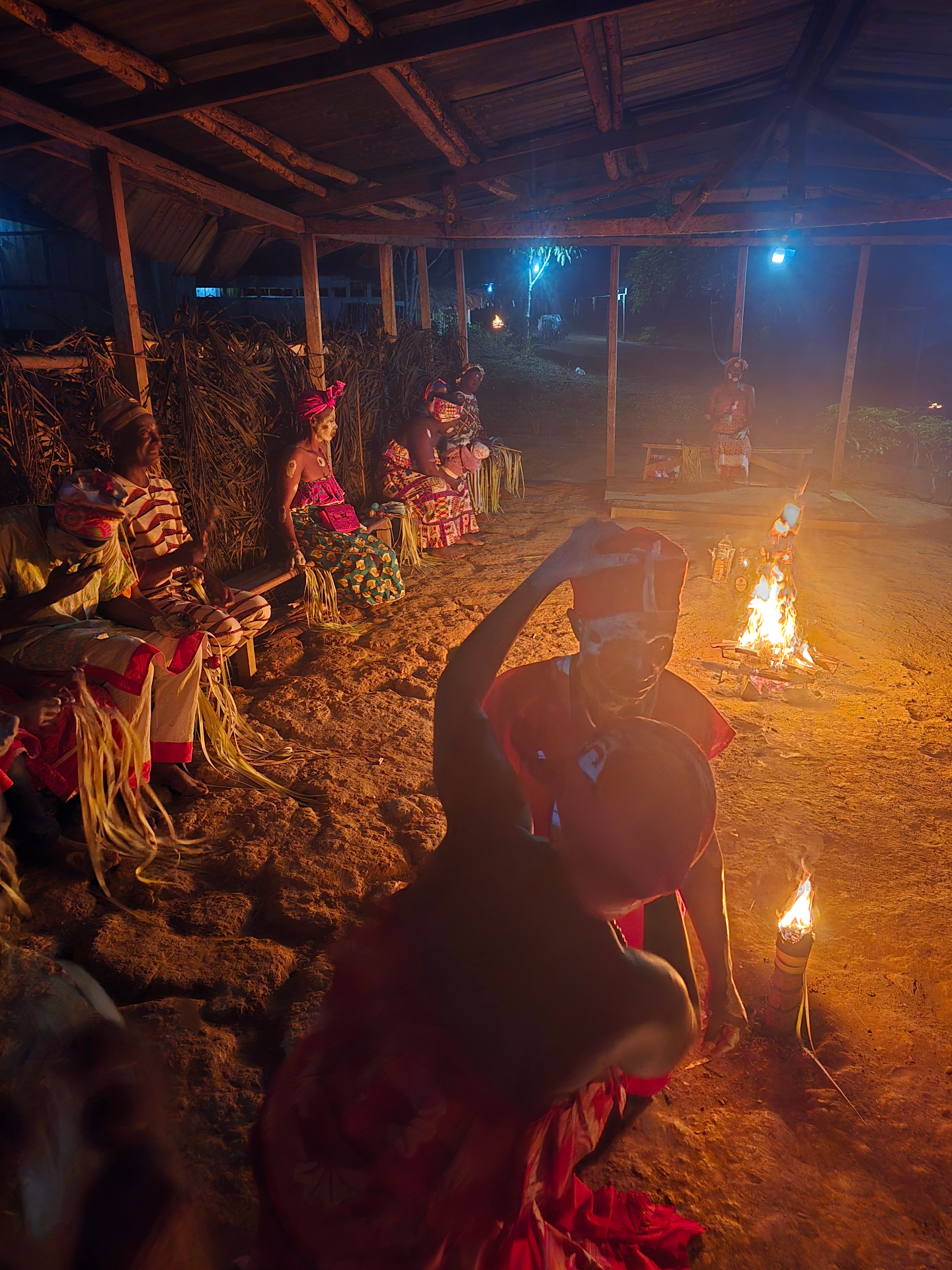 A nighttime Bwiti ceremony at Ntann, initiates seated under a thatched temple as a fire burns on the sand.